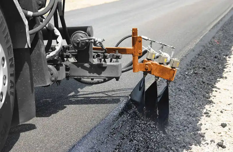 Bitumen emulsion CMS applied along the edge of a newly paved road with specialized asphalt sprayer.